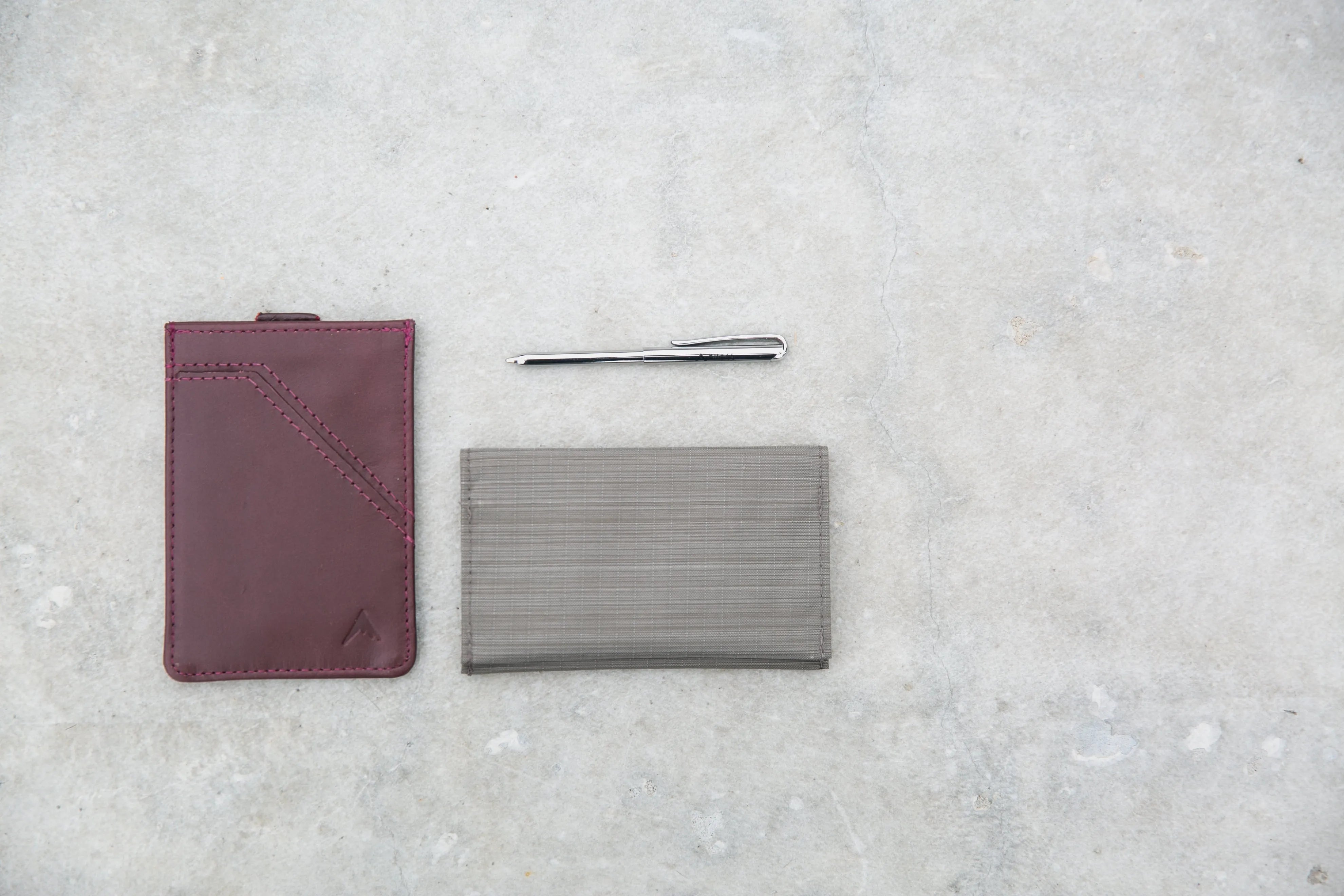 On the left is a maroon leather wallet. On the right is a light grey vegan nylon wallet. Above the vegan wallet is a micropen.