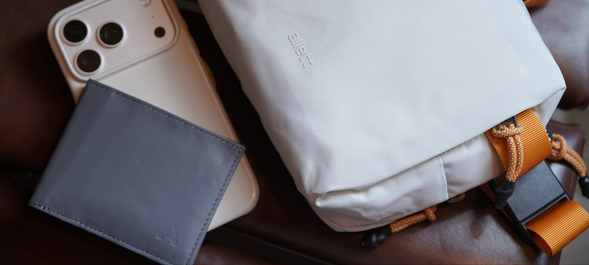 Allett lunar ash nylon sling bag with orange straps and hardware, styled on a dark brown leather surface next to a slate gray wallet and a light-colored smartphone.