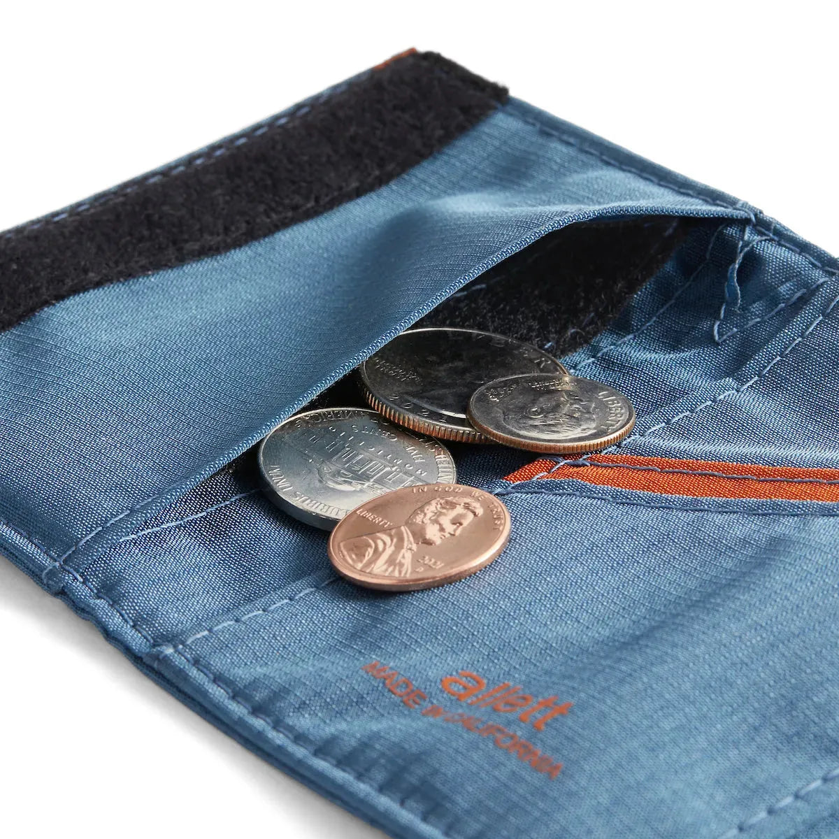 An assortment of coins spilling from the interior coin holder beneath the velcro side pocket of the indigo blue trifold wallet from Allett. Allett Trifold Wallet nylon edition - indigo-blue