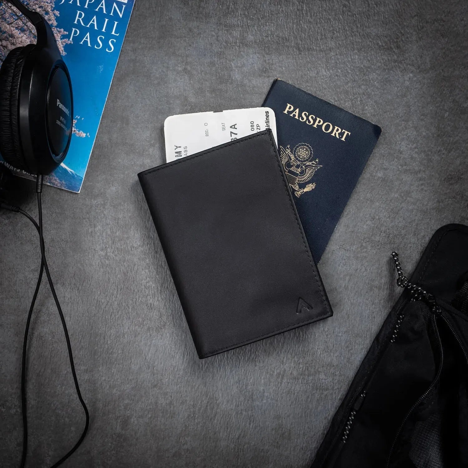 Black leather passport wallet by Allett against a grey table. Underneath the wallet is a passport, and there is also headphones on the table - black