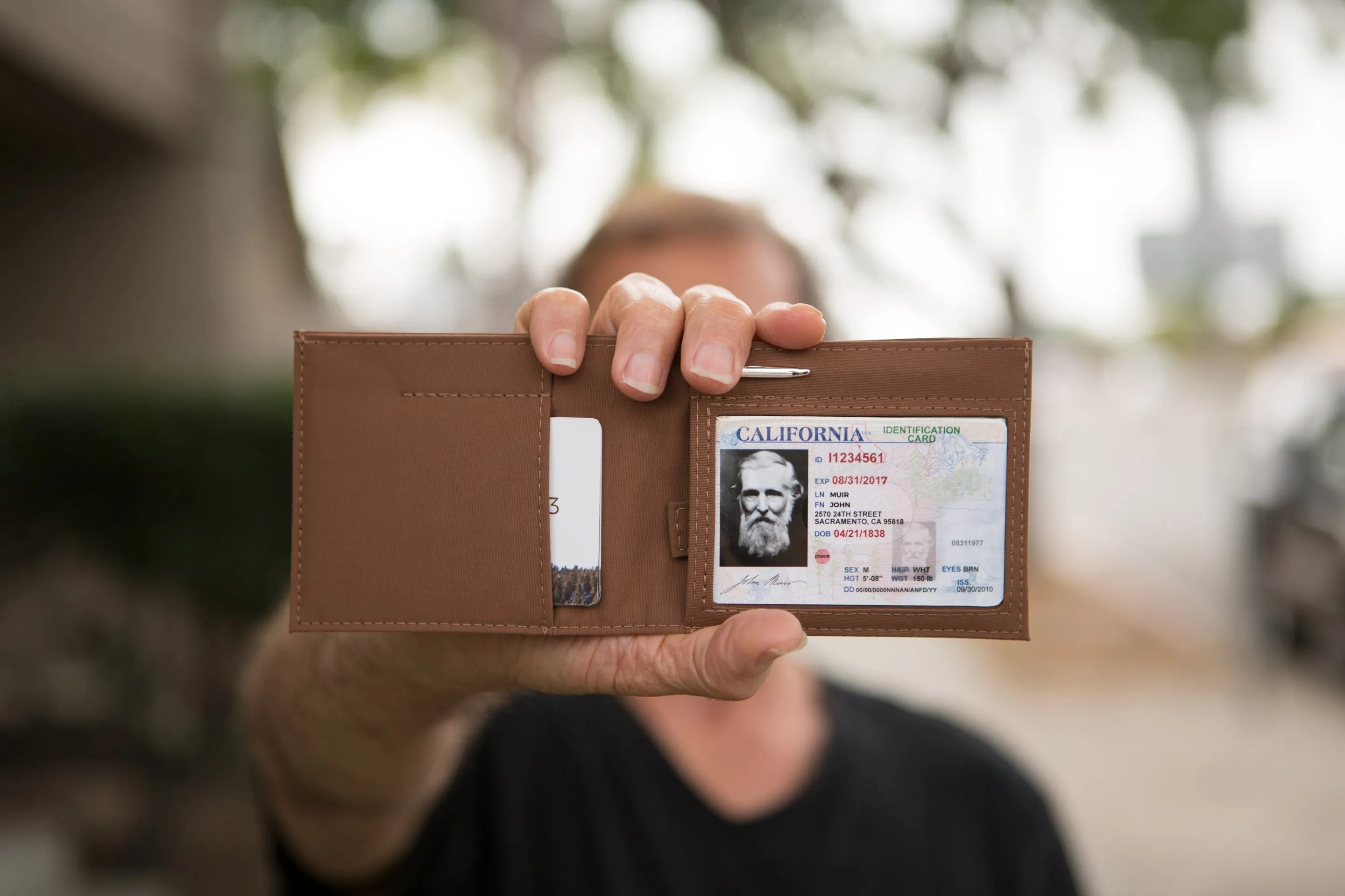 Unnamed man holding a brown leather wallet in front of him with a viewable sample identification card.