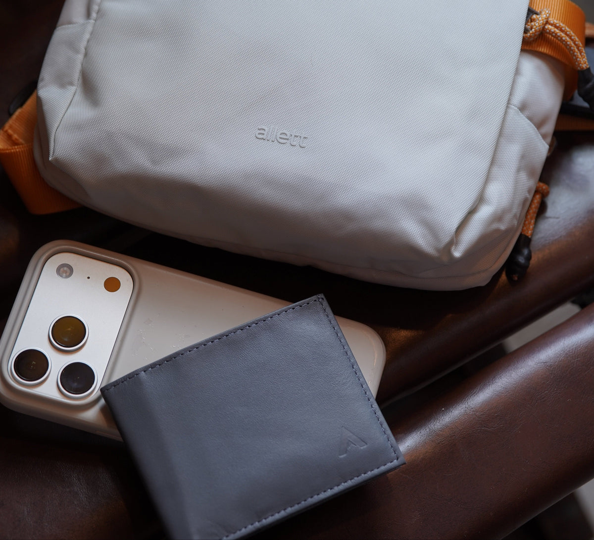 Allett lunar ash sling bag with orange- strap and a small gray leather cardholder, shown with a smartphone in a white case resting on a brown leather surface.